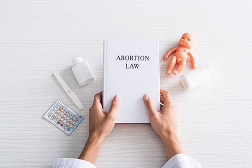 top view of woman holding book with abortion lettering near baby doll and abortion pills