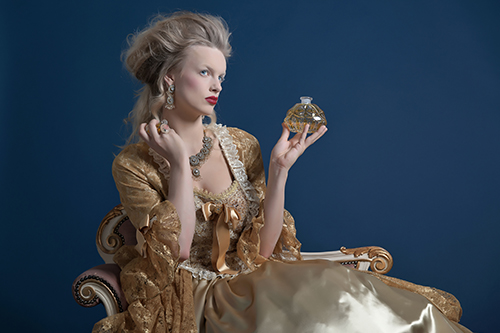 Retro baroque fashion woman wearing gold dress. Holding bottle of parfume. Sitting on vintage couch. Studio shot against blue.
